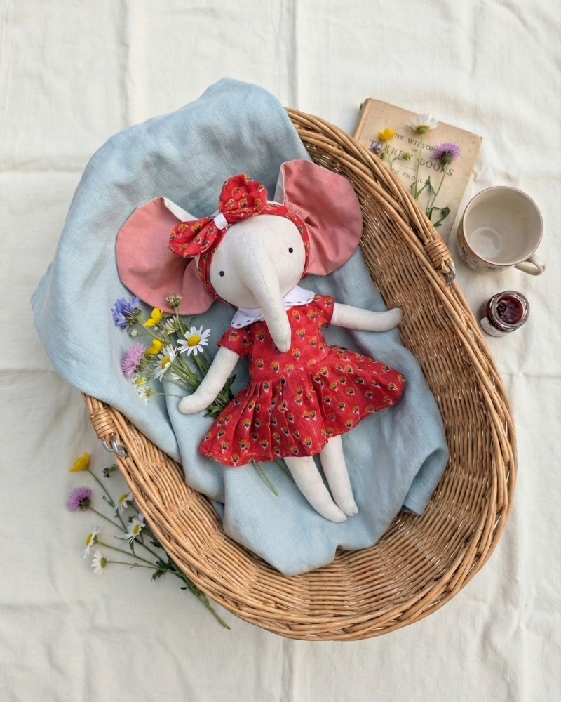 Elephant soft doll in a red dress with a red bow on a light blue blanket in a wicker basket.