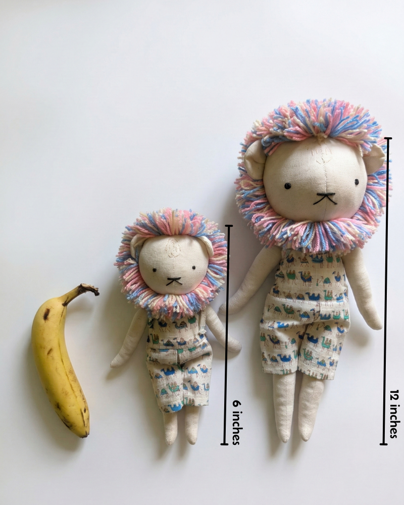 Two lion-shaped toys with colorful manes next to a banana on a white background, with measurements indicated.